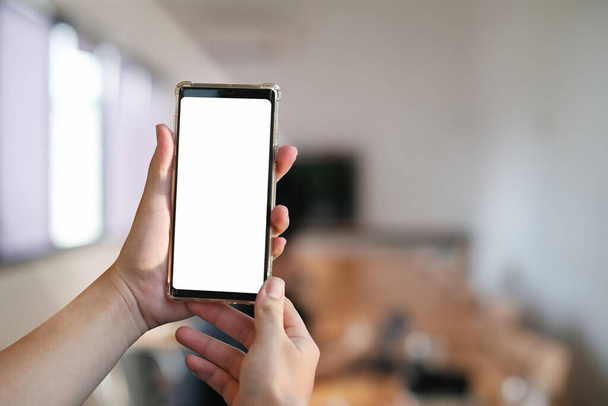Обрезанный снимок рук, держащих смартфон с чистым экраном и размытым офисным фоном. - Фото, изображение