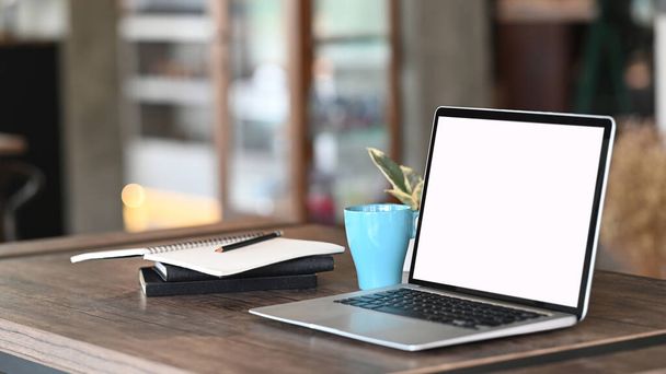 Computer-Laptop mit weißem Bildschirm, Kaffeetasse und Notizbuch auf Arbeitstisch vor verschwommenem Wohnzimmer-Hintergrund. - Foto, Bild