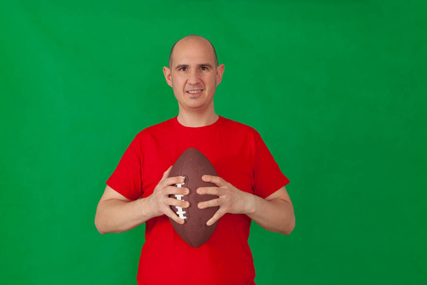 A bald caucasian man dressed in a red t-shirt holding a soccer with both hands in front of his chest. The background is green. - Fotografie, Obrázek