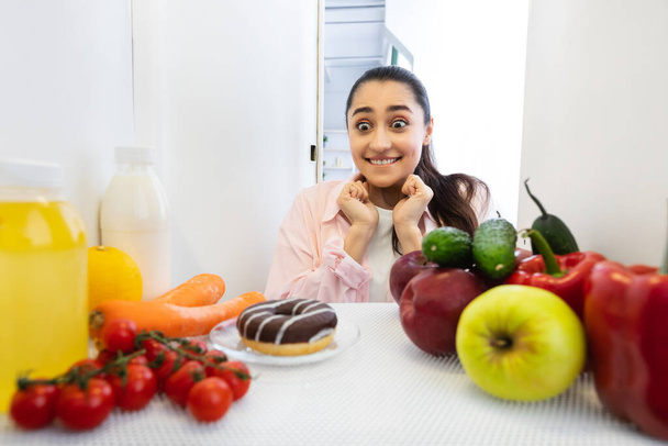 Aufgeregt lustige Frau schaut auf leckeren Donut Blick aus Kühlschrank - Foto, Bild