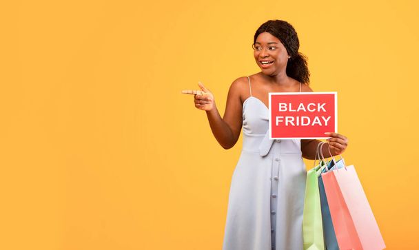Encantadora mujer negra con bolsas de comprador sosteniendo el letrero BLACK VIERNES y señalando a un lado el espacio vacío, fondo naranja - Foto, imagen