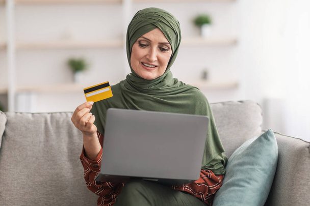 Maduro musulmán mujer compras usando portátil y tarjeta de crédito interior - Foto, imagen