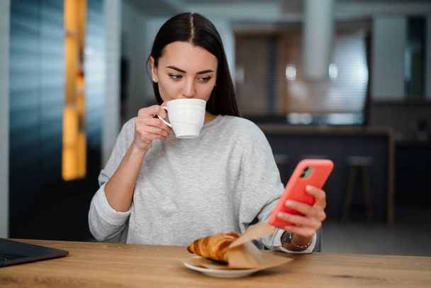 Focused young woman using mobile phone while drinking coffee with croissant at home - Foto, Imagem