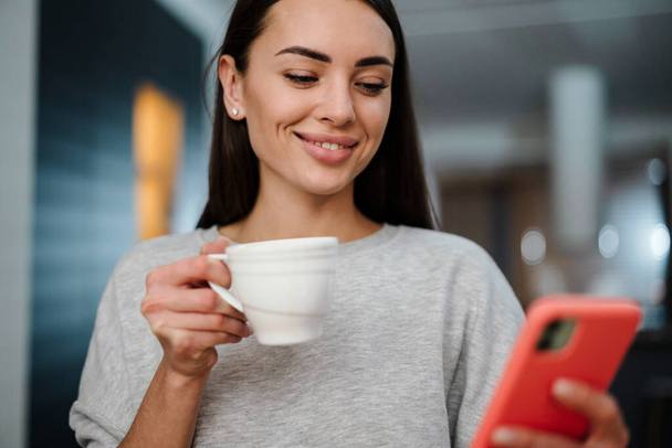 Happy young woman using mobile phone while drinking coffee at home - Foto, Bild