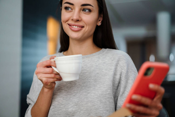 Happy young woman using mobile phone while drinking coffee at home - Foto, Imagen
