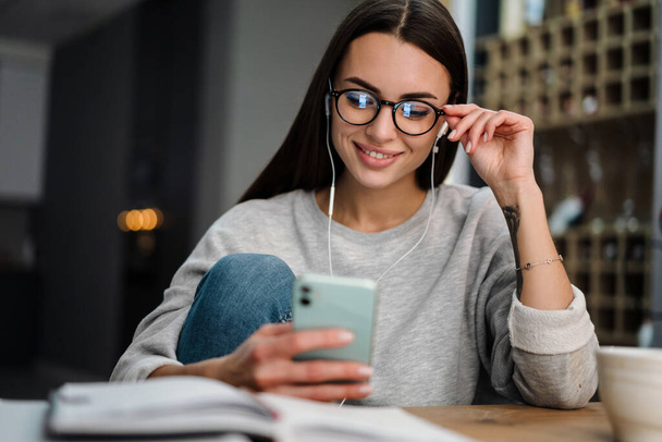 Happy young woman listening music with earphones and mobile phone while sitting at home - Zdjęcie, obraz