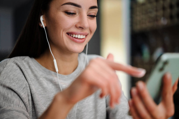 Happy young woman listening music with earphones and mobile phone while sitting at home - Фото, изображение
