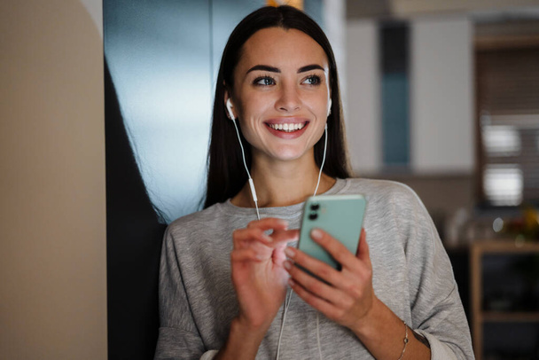 Smiling young woman listening music with earphones and mobile phone at home - Foto, Imagen