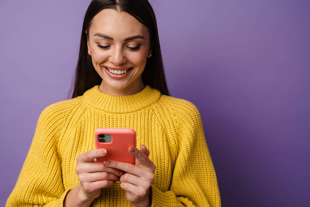 Happy young woman wearing sweater holding mobile phone over violet background - Fotografie, Obrázek