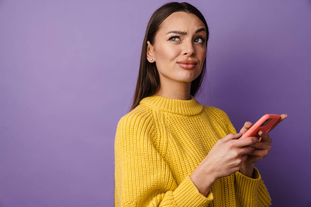 Pensive young woman wearing sweater holding mobile phone over violet background, looking up - Fotografie, Obrázek