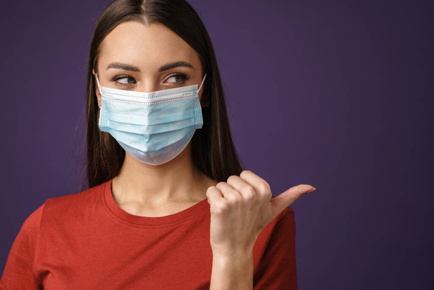 Beautiful brunette girl in protective mask pointing finger aside isolated over purple background - Photo, Image