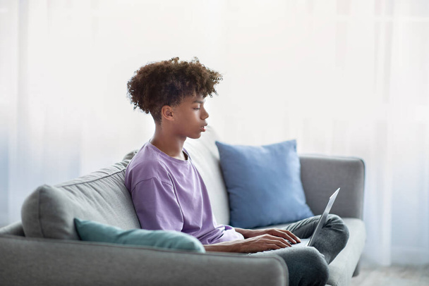 Η ιδέα της εξ αποστάσεως εκπαίδευσης. Μαύρος έφηβος που χρησιμοποιεί φορητό υπολογιστή για τις online σπουδές του στο σπίτι, πλαϊνή όψη - Φωτογραφία, εικόνα