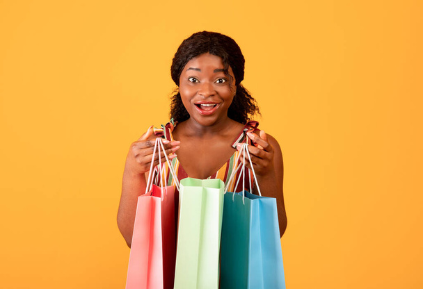 Letnia koncepcja zakupów. Zaskoczona czarna kobieta z papierowymi torbami patrząca na kamerę na pomarańczowym tle studia - Zdjęcie, obraz