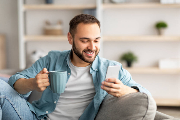 Positive young guy holding smartphone and coffee indoors, chatting with friends online, sending email, browsing internet - Foto, Bild