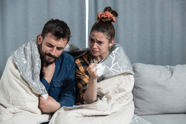 Young couple suffering from a common cold and flu sitting at home wrapped in blankets and wipe their noses with tissues while they have a strong headache, healthcare concept - Photo, image