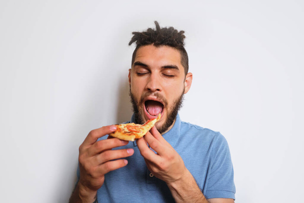 Joven lindo hombre caucásico con barba y rastas come pizza sobre fondo blanco neutro. Humano sostiene un pedazo de pizza de queso con tomates en sus manos y disfruta comiendo. Banner horizontal. - Foto, imagen