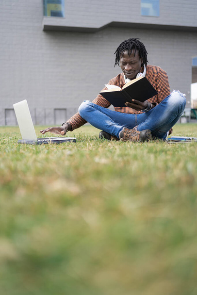 Estudiante Africano leyendo libro y utilizando el portátil para investigar información para el proyecto de la Universidad, al aire libre en la hierba del campus - Foto, imagen