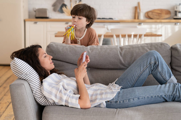 Calm mom lying on sofa with smartphone distracted from noisy loud son bad behavior drawing attention - Φωτογραφία, εικόνα