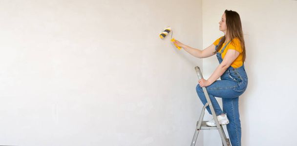 A pensive woman with roller and ladder looks dreamily in a room with an unpainted wall with copy space. Independent single female makes DIY repairs in her apartment - Foto, Imagen
