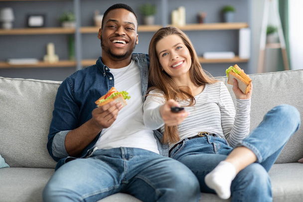 Couples Leisure. Cheerful Young Interracial Lovers Watching Tv And Eating Sandwiches - Fotoğraf, Görsel