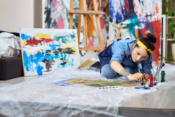 絵の具の色を選ぶエプロンの才能ある女性画家、絵を描く作業中に絵筆を持ち、自宅のワークショップで床に座って - 写真・画像