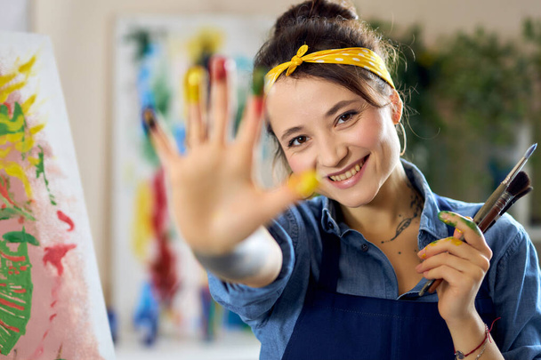 Artiste féminine joyeuse montrant sa main dans la peinture à la caméra tout en créant la peinture, l'application de peintures colorées sur toile avec les doigts - Photo, image
