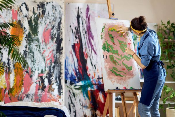 Back view of talented female artist working on a modern abstract oil painting. She is applying paint on canvas with fingers, standing in home studio workshop - Photo, Image