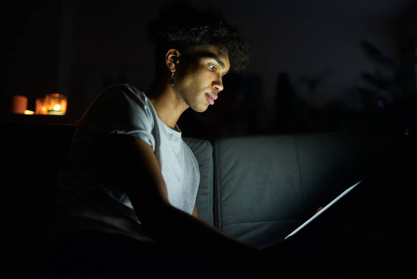 Side view of focused young guy reading e book or watching video using tablet pc at night, sitting in dark room at home - Фото, изображение