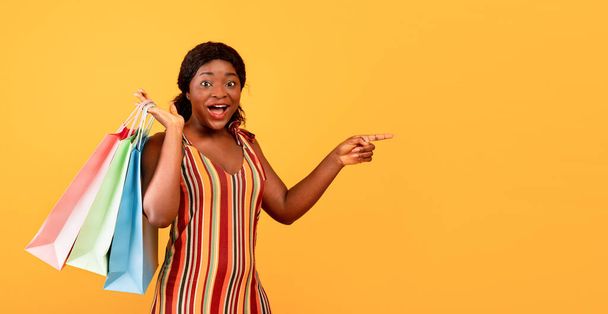 Excited black lady in dress holding shopper bags, cannot believe big sale, pointing at empty space on orange background - Fotoğraf, Görsel