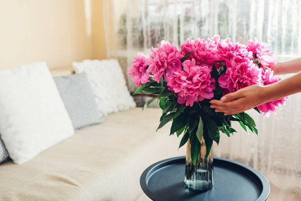 Woman holding bouquet of peonies in vase at home. Pink blooms put on coffee table. Interior and summer decor - Foto, Imagen