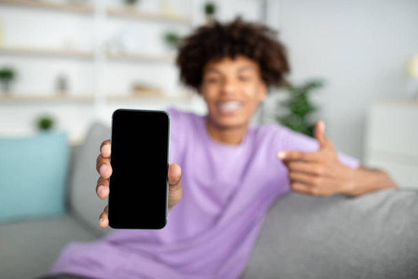 Cool African American nastolatek wskazując na telefon komórkowy z pustym ekranem w domu, makieta dla aplikacji mobilnej lub stronie internetowej - Zdjęcie, obraz