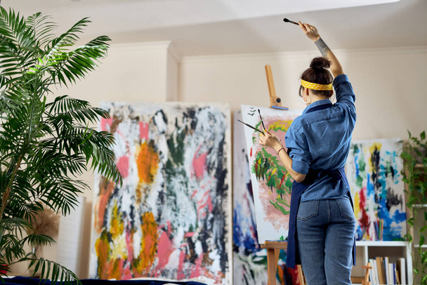 Pohled zezadu ženské umělkyně zvedla ruku s štětcem a vytvořila novou malbu na plátně, stojící v dílně domácího studia - Fotografie, Obrázek