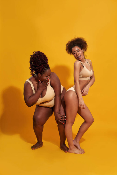Two young african american women in underwear with different body shape having fun while standing together isolated over yellow background - Foto, Bild