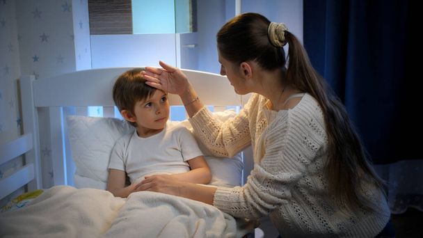 Fiatal anya ül a beteg kisfia mellett, aki éjszaka fekszik az ágyban. A gyermekbetegség, a betegség és a szülői ellátás fogalma - Fotó, kép