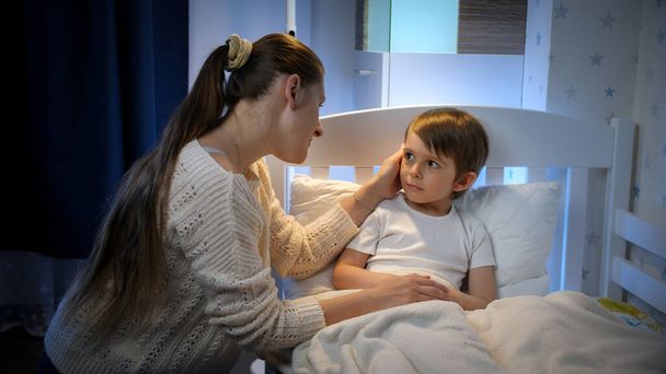 Fiatal gondoskodó anya megérinti a beteg kisfia homlokát az ágyban fekve. A gyermekbetegség, a betegség és a szülői ellátás fogalma. - Fotó, kép