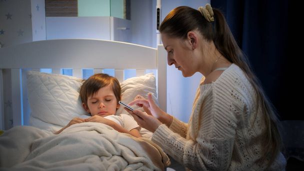 Junge Mutter sitzt neben ihrem kranken Kind im Bett und sucht mit dem Smartphone nach Medikamenten. Konzept der Kinderkrankheit und der elterlichen Betreuung - Foto, Bild
