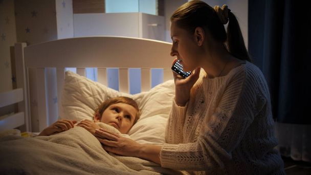 Mały chory chłopiec leżący w łóżku obok matki rozmawiający przez telefon z lekarzem. Pojęcie choroby dzieci, choroby i opieki rodzicielskiej - Zdjęcie, obraz