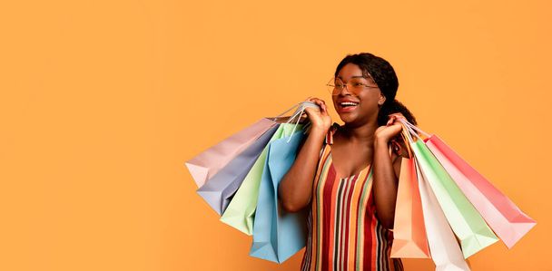 Attraktive junge schwarze Frau in Sommerkleidung hält Geschenktüten in der Hand und betrachtet die Leerstelle vor orangefarbenem Studiohintergrund - Foto, Bild