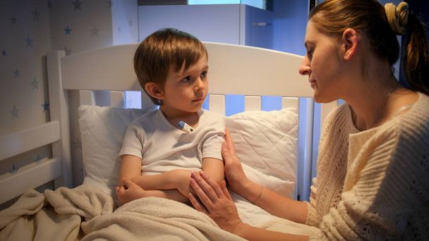Mały chory chłopiec mierzący temperaturę ciała w nocy i patrzący na siedzącą obok młodą matkę. Pojęcie choroby dzieci, choroby i opieki rodzicielskiej - Zdjęcie, obraz