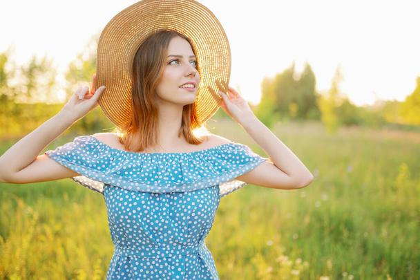 Nuori nainen pellolla auringonlaskun aikaan säätää hattua ja hymyilee. - Valokuva, kuva