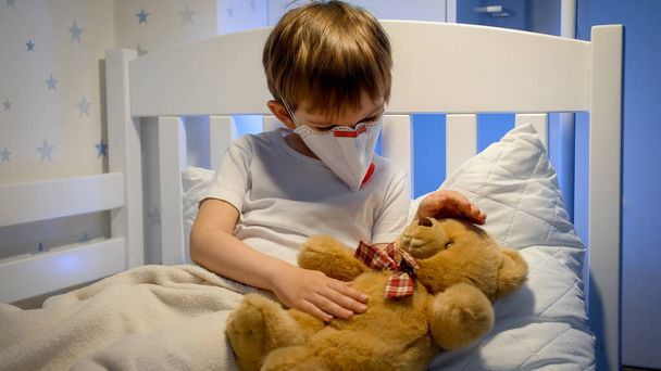 Kleine jongen draagt een beschermend medisch masker in bed en speelt met teddybeer. Concept van het kind virus en kinderen bescherming tijdens coronavirus Covid-19 pandemie. - Foto, afbeelding