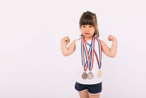 Маленькая девочка с каштановыми волосами с медалями чемпиона спорта на шее делает символ силы. Концепция спорта и победы - Фото, изображение