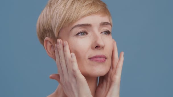 Semi profile portrait of middle aged blonde lady massaging face and neck, applying lotion, blue background, slow motion - Footage, Video