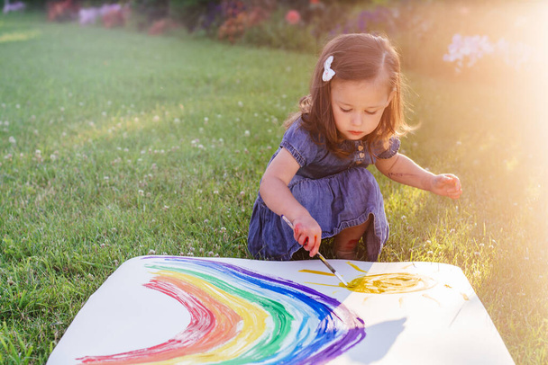 Κοριτσάκι 2-4 ετών ζωγραφίζει ουράνιο τόξο και ήλιο σε μεγάλο φύλλο χαρτιού που κάθεται στο πράσινο γκαζόν στο φως του ήλιου - Φωτογραφία, εικόνα