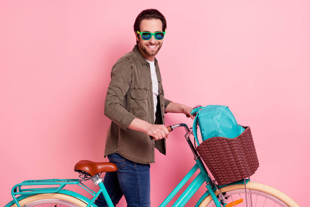 Foto von entzückenden süßen jungen Kerl gekleidet braunes Hemd dunkle Brille Reiten Fahrrad Rucksack isoliert rosa Farbe Hintergrund - Foto, Bild