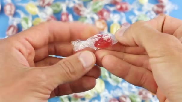 le mani spiegano un lecca-lecca di caramelle rosse da un involucro trasparente, sullo sfondo di molte caramelle colorate, caramelle malsane, dipendenza da zucchero - Filmati, video