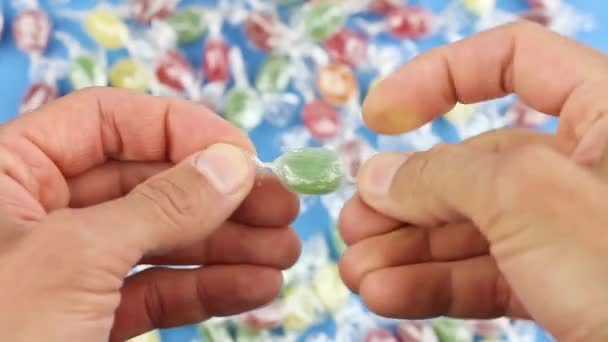 las manos despliegan una piruleta de caramelo verde de una envoltura transparente, sobre el fondo de muchos caramelos de colores, dulces de azúcar poco saludables, adicción al azúcar - Imágenes, Vídeo