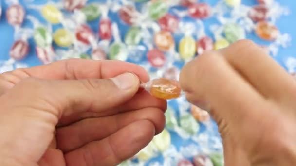 hands unfold one orange candy from a transparent wrapper, on background of many colored candies, sugar sweets - Footage, Video