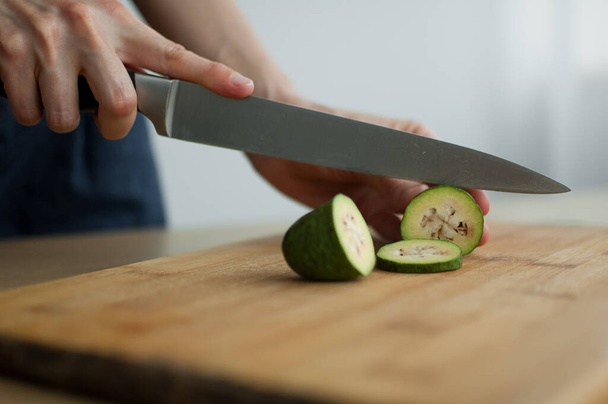 Las manos femeninas están cortando una fruta feijoa verde fresca en una tabla de madera cortada. Frutas exóticas, concepto de alimentación saludable - Foto, Imagen