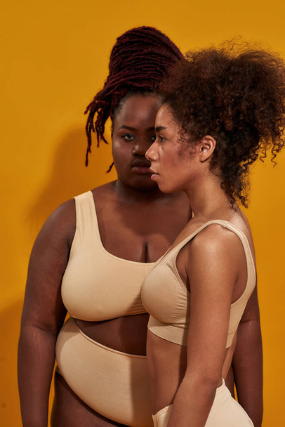 Front and side view of two african american female models in underwear with different body size, standing together isolated over yellow background - Фото, изображение